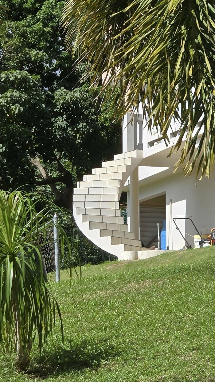 Spiral death stairs outdoors, against a white building, surrounded by greenery and trees.