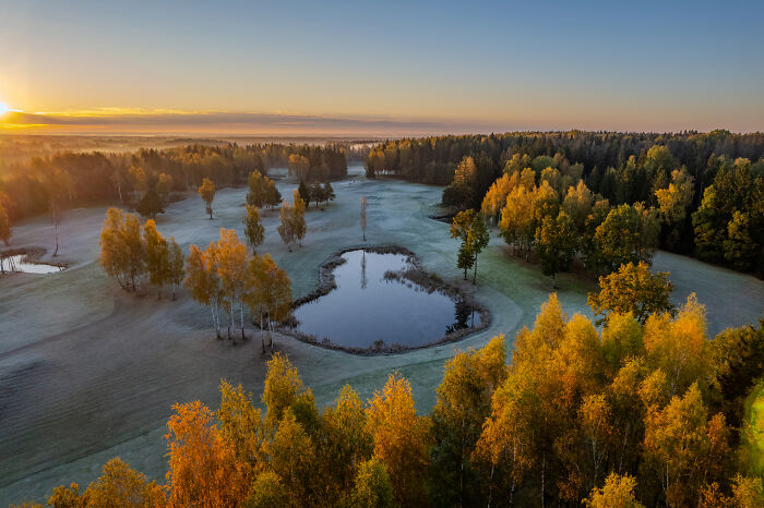 Bird’s-eye view of a Lithuanian landscape with a pond, surrounded by autumn trees at sunrise.