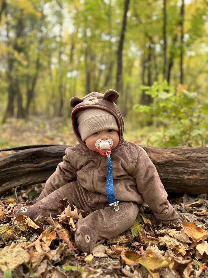 Baby dressed in bear costume sitting among autumn leaves in forest, capturing wholesome parenting moments with dads.