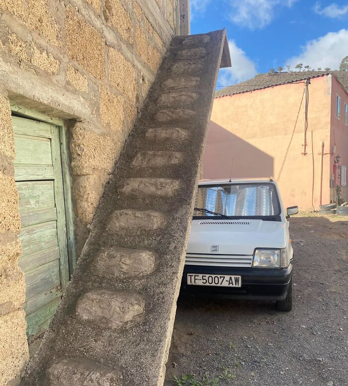 Unusual steep stairs leaning against a wall, with a car parked beneath. Death stairs concept depicted outdoors.