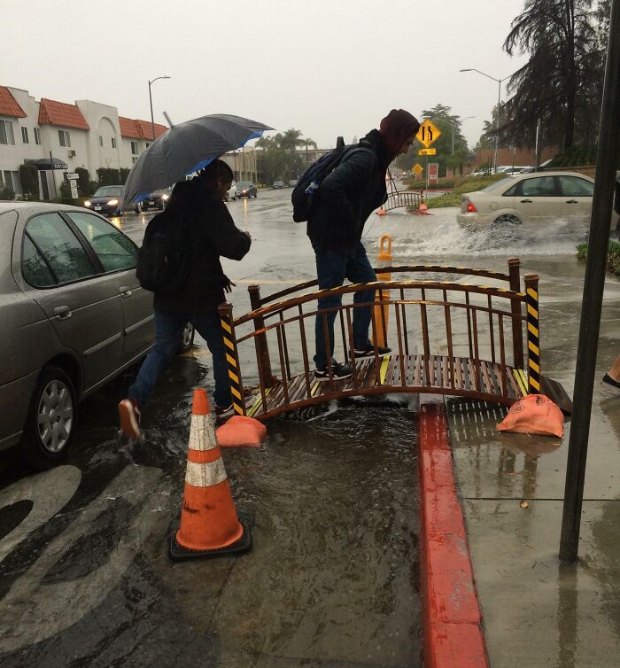 People using a small bridge to cross a flooded street, illustrating cities genius solutions in urban design during rainstorms.