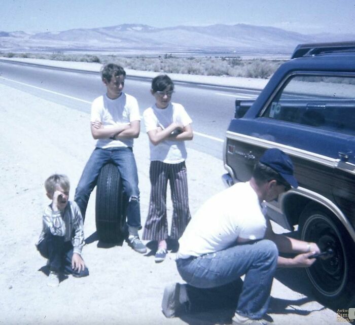 Three children watch as a man changes a car tire on a desert roadside in an awkward family photo moment.