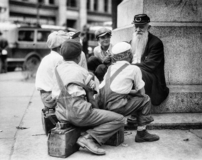 Children in vintage attire sitting with an elderly man, showcasing something interesting from the past.