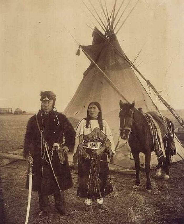 Two individuals in traditional attire with a horse, standing in front of a teepee, showcasing something interesting.