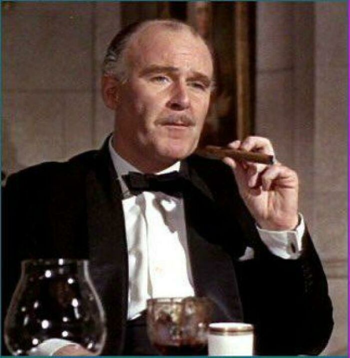 Man in a tuxedo holding a cigar, seated at a dining table.