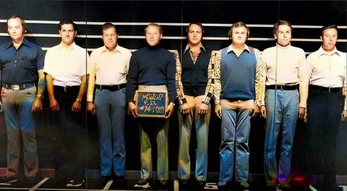 Seven men standing in a police lineup, each dressed in 1970s attire.