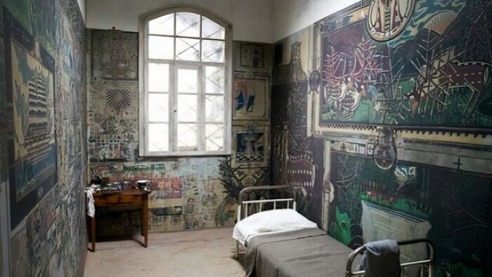 Room with intricate wall art, featuring a window, a bed, and a small table, showing something interesting.