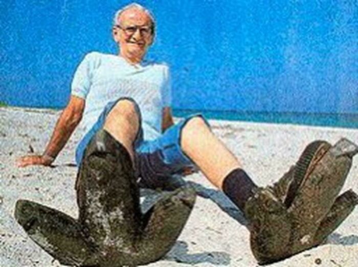 Elderly man sitting on a beach wearing extremely oversized boots, showcasing strange Earth phenomena in a surreal scene.