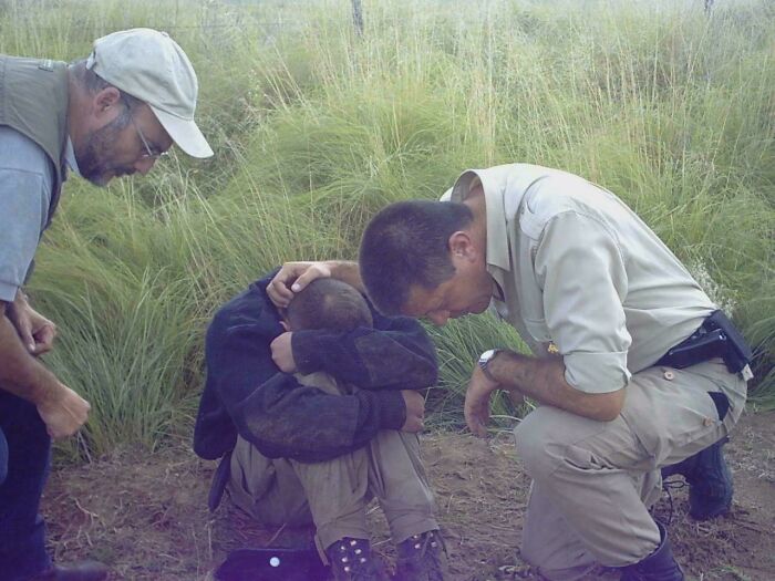 A distressed person sitting on the ground in tall grass while two men in outdoor gear offer support, strange earth scene.