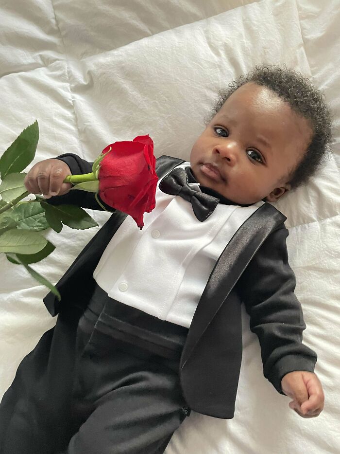 Baby dressed in a tuxedo holding a red rose, capturing a wholesome parenting moment with dads doing their best.