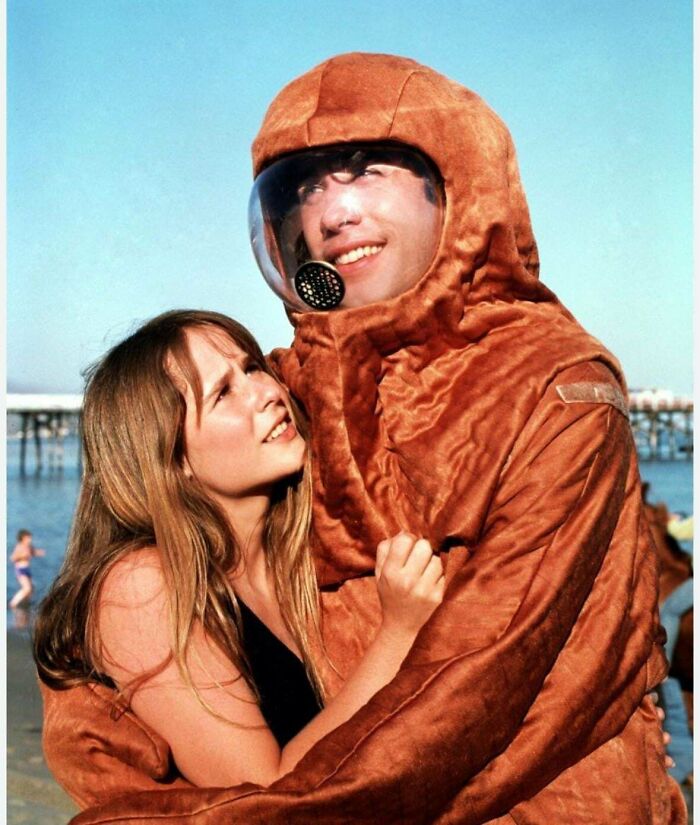 A woman embraces a man in a vintage brown spacesuit on the beach under a clear blue sky, evoking nostalgia.