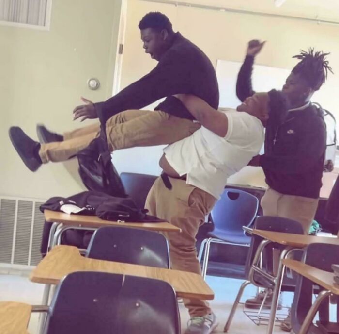 Three students in a classroom engaged in playful lifting, creating an accidental Renaissance painting effect.