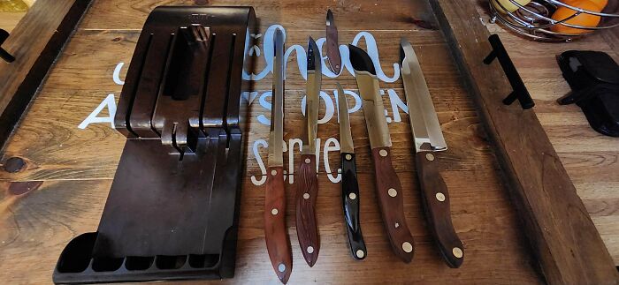 A Buy It For Life knife block and set of various kitchen knives with wooden and black handles, on a rustic wood surface.