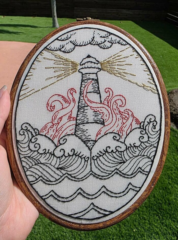 Hand holding embroidery hoop with incredible lighthouse design and detailed waves showcasing beautiful embroidery skills.