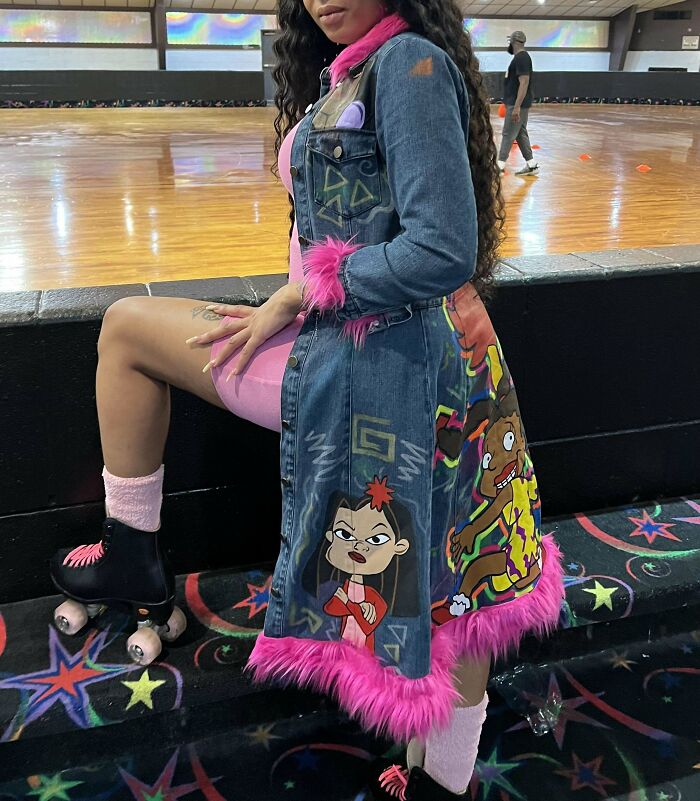 Person in vibrant DIY roller skating outfit with cartoon graphics and pink fur trim, sitting on rink edge.