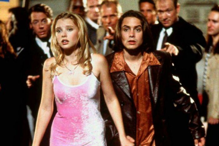 Two people in 2000s fashion, a woman in a pink dress and a man in a brown leather jacket, walking through a crowd.