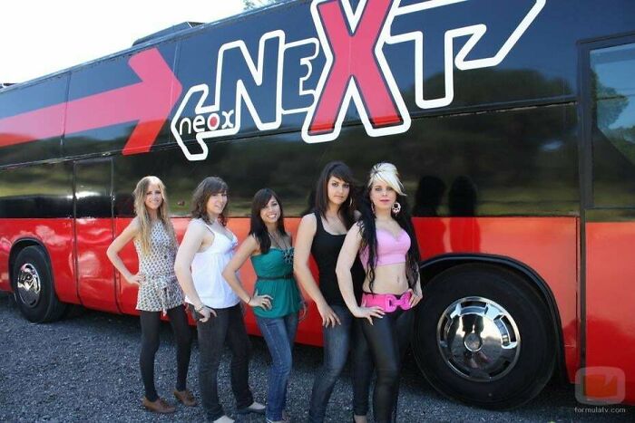 Five women posing in front of a colorful 2000s-themed tour bus labeled "Next."
