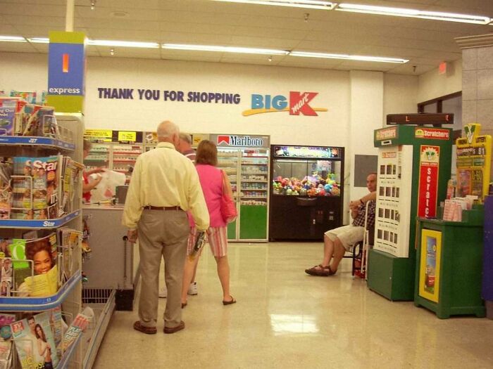 Shoppers in a 2000s Kmart store interior with checkout, magazines, and vending machines, evoking nostalgia.