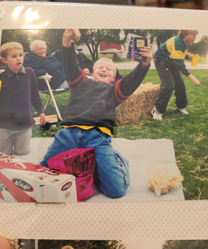 Child excitedly holding a Game Boy Color in a 2000s-themed outdoor gathering with family and friends.