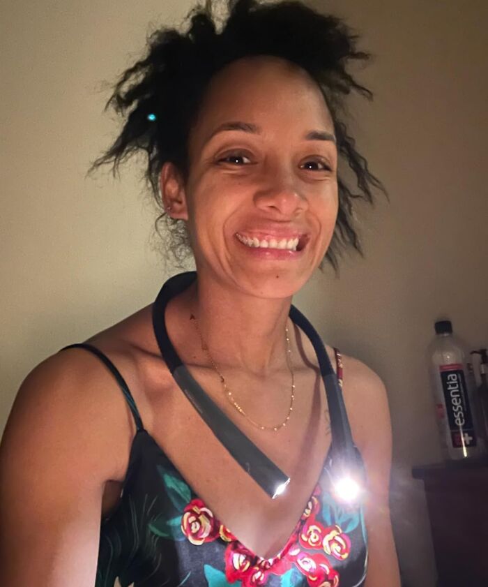 Woman wearing a clever invention: a neck light, smiling indoors by a table with a bottle.