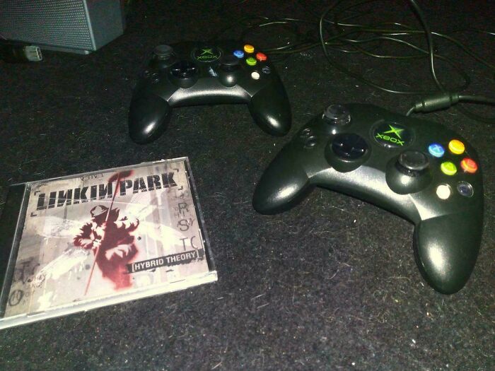 Two Xbox controllers and a Linkin Park CD from the 2000s on a dark surface.