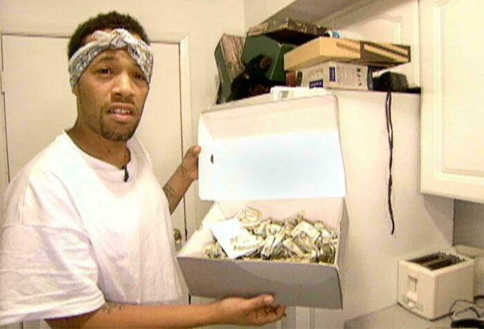 Man holding a shoebox filled with money in a 2000s-style kitchen, wearing a white shirt and bandana.