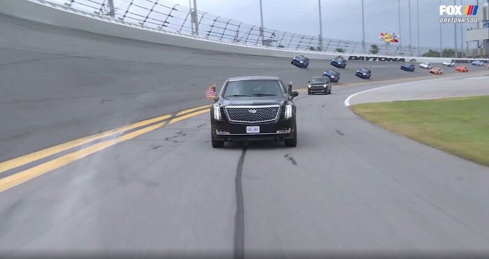A black SUV leading a line of race cars on a track, with an American flag, during a race; facepalm moment.