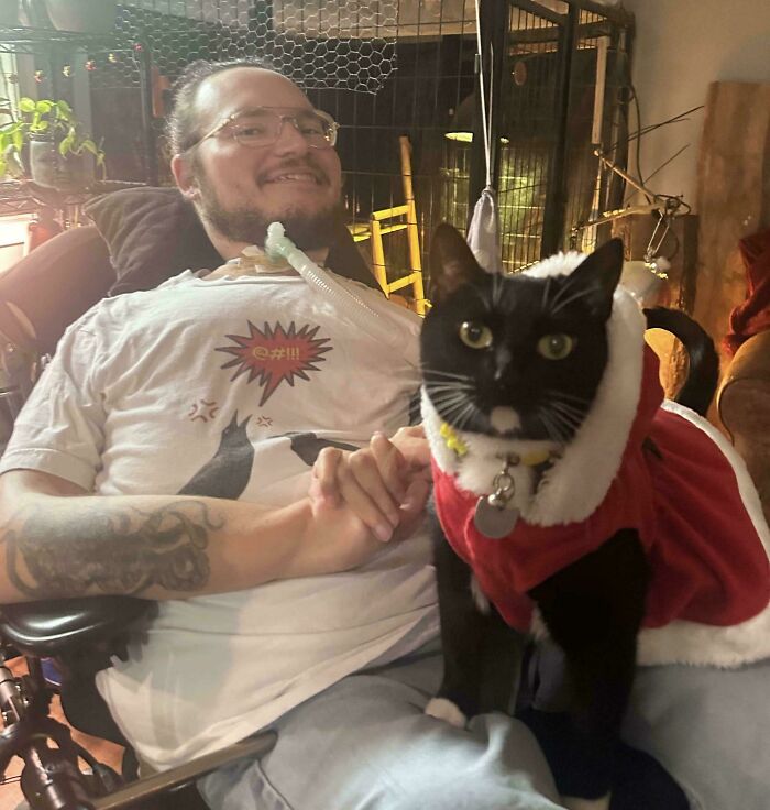A tuxedo cat wearing a festive outfit sits next to a smiling person in a wheelchair.