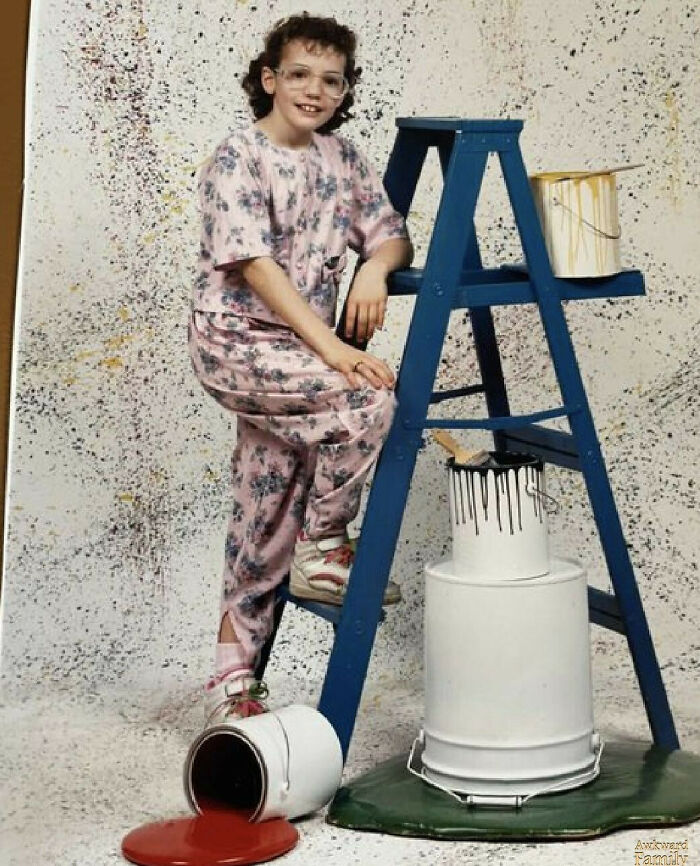 Young girl in floral outfit posing awkwardly on a ladder with spilled paint, capturing an awkward family photo with sweet charm.