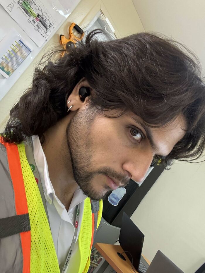 Man with fierceflow hair style wearing safety vest and earbuds in an office setting with computers and documents on walls.