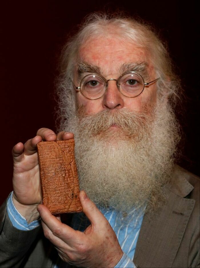 Elderly man with glasses and a long beard holding an ancient artefact, a carved clay tablet, against a dark background.