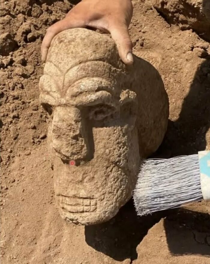 Ancient artefact being unearthed, a carved stone head cleaned with a brush, showcasing remarkable preservation.