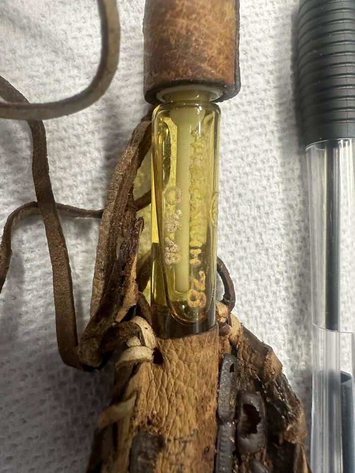 Weird object with a leather strap and a glass vial containing yellow liquid, next to a black pen.