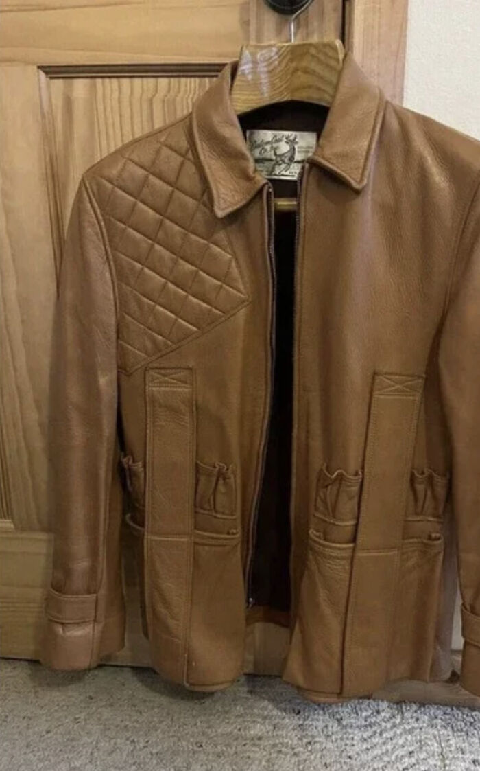 Brown leather jacket with quilted shoulder and unusual pocket design, hanging on a wooden hanger.
