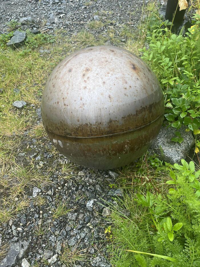 Rusty spherical object on grass and rocky ground, resembling a large metal ball. Weird object in nature.