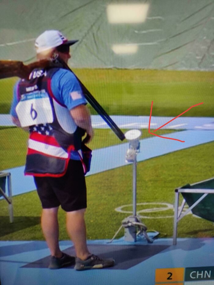 Person holding a rifle next to a mysterious object marked with an arrow, featuring a flag-themed vest and a cap.