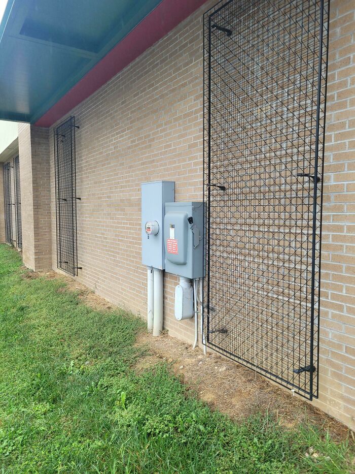 Weird objects: Electrical meter with wiring on a brick wall next to metal grate panels.