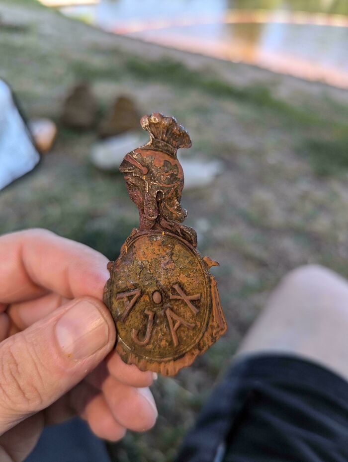 Hand holding a rusty, mysterious object labeled "AJAX" with a sculpted figure on top, found during an outdoor exploration.