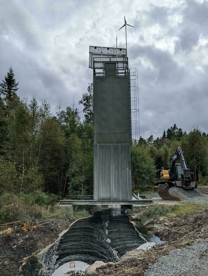 Weird object resembling a tall metal structure with ladder, small wind turbine on top, near forest and construction equipment.