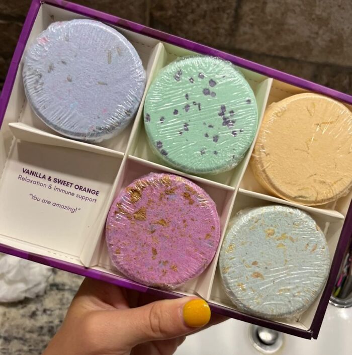 Colorful bath bombs in a gift box, labeled vanilla and sweet orange, held by someone with yellow-painted nails.