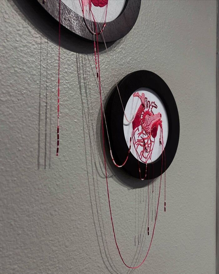 Framed embroidery of an anatomical heart with delicate red threads hanging, showcasing incredible embroidery skills.