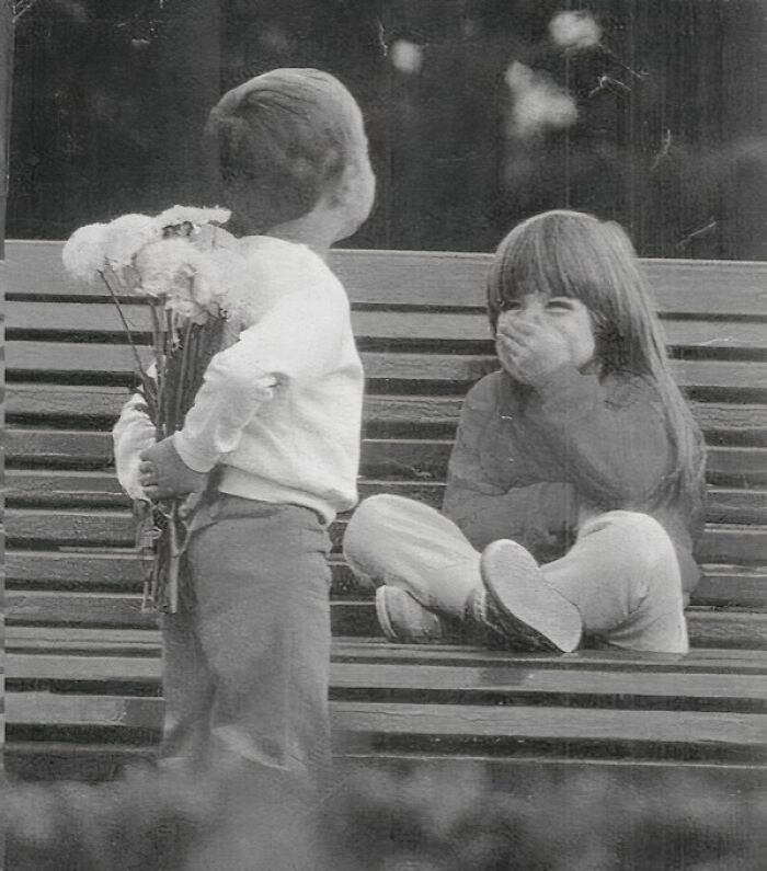 Two children on a bench, boy hiding flowers behind his back, girl giggling, capturing true happiness.