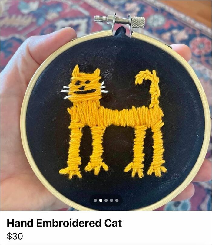 Hand holding a humorous DIY embroidery of a cat, stitched in yellow thread on a black fabric background.