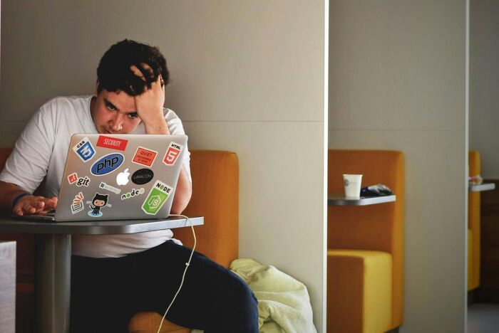 Man facing work-struggles, sitting in a booth, hand on head, looking at a sticker-covered laptop.
