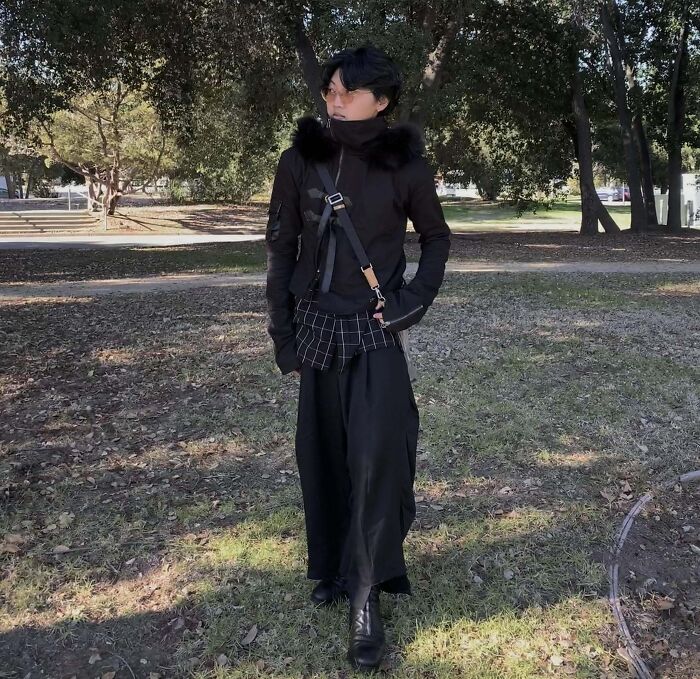 Person showcasing a bold fashion look with a black outfit, including a fur-collared jacket and wide-legged pants, in a park setting.