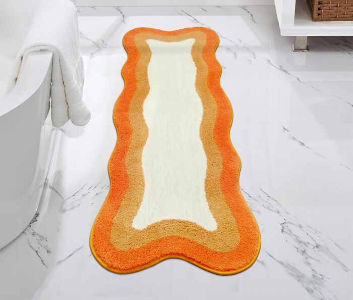 Wavy orange and beige bathroom mat on white marble floor next to a bathtub with towels hanging over edge