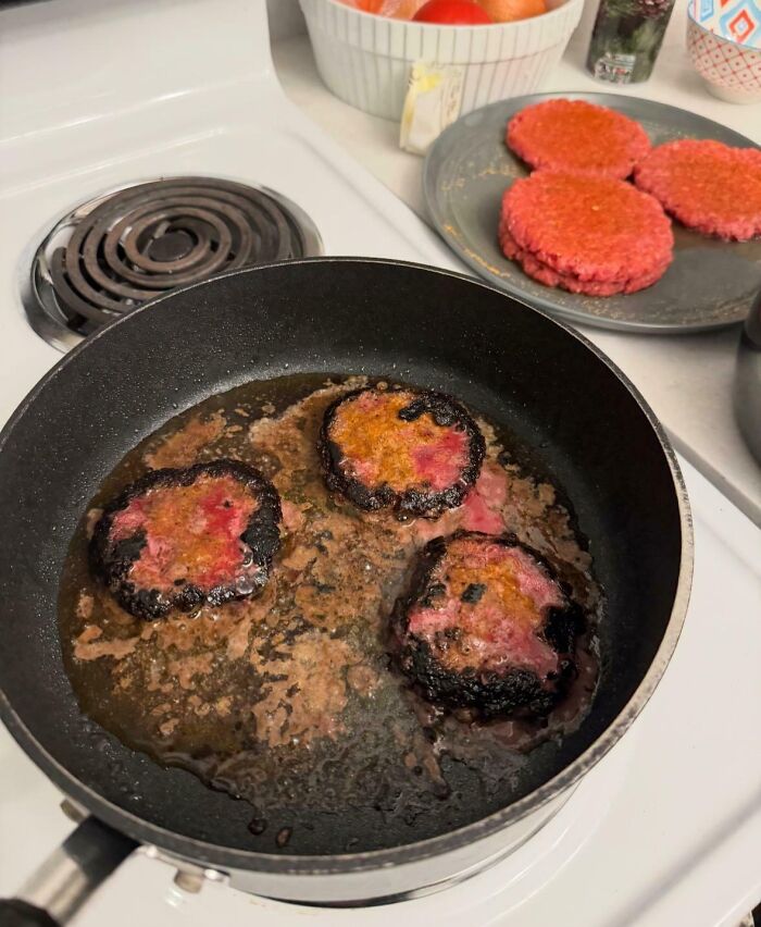 Burnt burgers in a frying pan near raw patties, illustrating a cooking mishap.