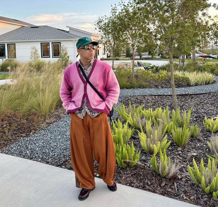 Person wearing a bold fashion outfit with a pink cardigan, printed shirt, orange trousers, and green cap.