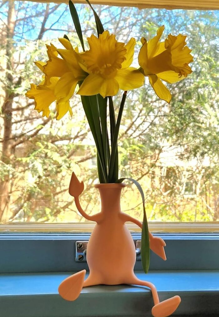 Playful vase shaped like a sitting figure holding yellow daffodils, on a windowsill.