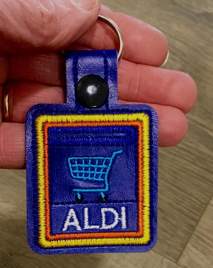 Close-up of a hand holding a blue ALDI keychain, featuring an embroidered shopping cart design.